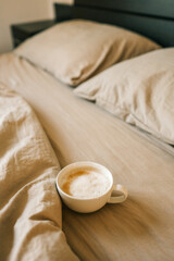White ceramic cup of frothy cappuccino resting on soft beige bedding. Cozy morning atmosphere with warm tones and natural light in a peaceful bedroom setting
