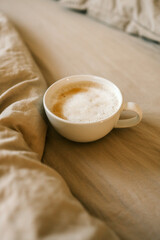 White ceramic cup of frothy cappuccino resting on soft beige bedding. Cozy morning atmosphere with warm tones and natural light in a peaceful bedroom setting