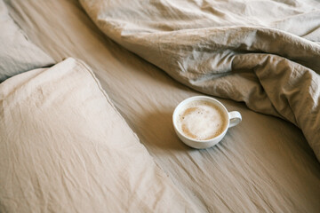 White ceramic cup of frothy cappuccino resting on soft beige bedding. Cozy morning atmosphere with warm tones and natural light in a peaceful bedroom setting