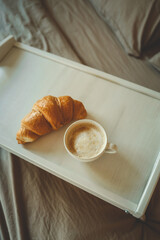 Morning breakfast in bed with fresh croissant and cappuccino on a white tray. Cozy neutral bedding and soft lighting create a relaxing start to the day