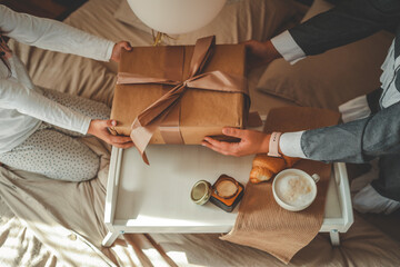 Child and adult exchanging a gift wrapped in kraft paper with ribbon on a cozy bed setup with breakfast tray, coffee, croissant, and candle. Soft morning light and warm tones create a festive mood