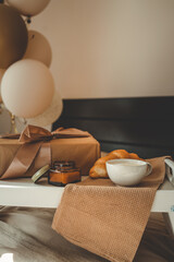 Breakfast in bed with a wrapped gift box, croissant, candle, and cup of coffee on a wooden tray, surrounded by soft bedding and festive balloons in beige and gold tones