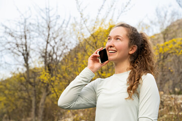 Fototapeta premium Young happy smiling woman with phone outdoors, talking on the smartphone, speaking on mobile cell phone in spring mountain park, resting in nature day during free time. Lifestyle concept