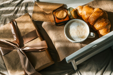 Breakfast in bed with a wrapped gift box, croissant, candle, and cup of coffee on a wooden tray, surrounded by soft bedding and festive balloons in beige and gold tones