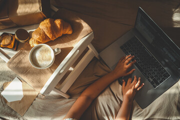 Child typing on a laptop on a bed beside a cozy breakfast tray with coffee, croissant, candle, and...