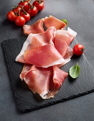  Italian prosciutto crudo on a black slate stone with cherry tomatoes on gray table. Generated image