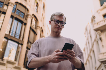 Young man in casual outfit looking at smartphone, standing between buildings on sunny street, using...
