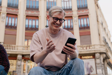 Casual young man enjoying relaxed afternoon in European city, holding smartphone while smiling and...