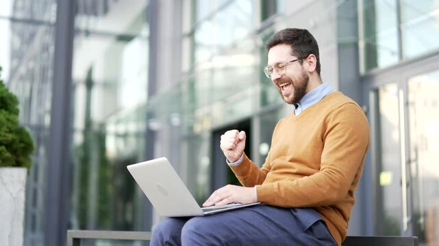 Happy excited businessman received great news on laptop while sitting on a bench on the street near office building. Smiling shocked surprised worker in glasses reads positive good message on computer