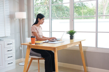 A casually dressed Asian businesswoman works from home while meeting online with a client, looking relaxed, focused and professional despite the informal setting.