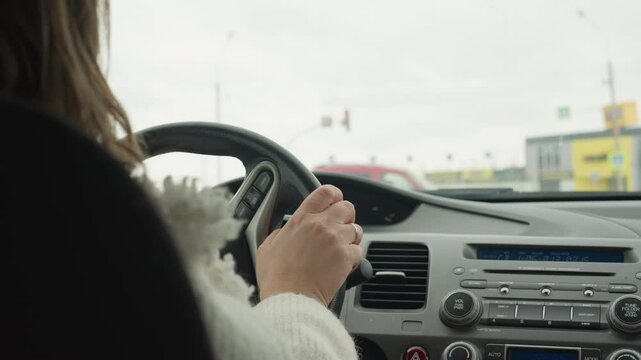 Back view of lady in fluffy white sweater holding steering wheel while patiently waiting at traffic light inside vehicle during daytime with blurred city view and passing cars seen through windshield