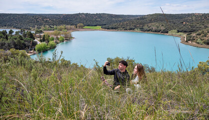 Young couple taking a selfie sitting on a grassy hillside with a scenic turquoise lake and rolling hills in the background under a partly cloudy sky