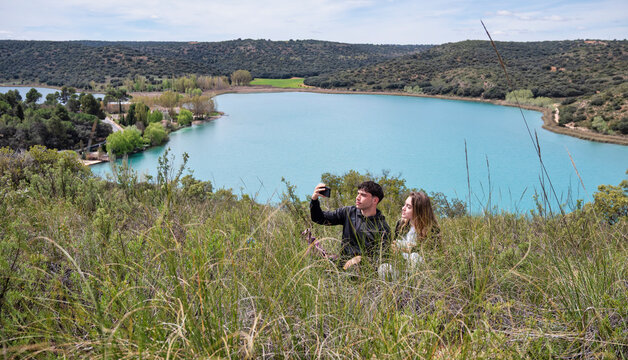 Young couple taking a selfie sitting on a grassy hillside with a scenic turquoise lake and rolling hills in the background under a partly cloudy sky - Powered by Adobe