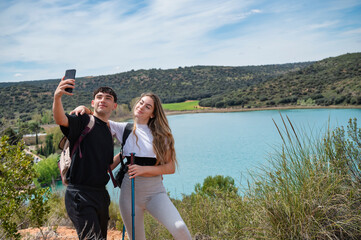 Teen couple taking a selfie during a hiking trip near a turquoise lake with forested hills and blue sky in the background