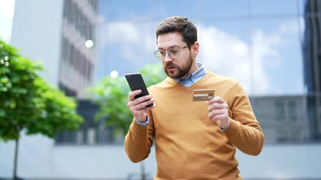 Frustrated businessman checks his credit card on phone standing on street near an business office building. Shocked worker faced fraud, money was stolen from his account. He became victim of deception