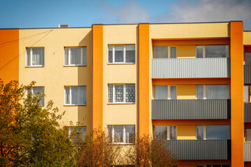 A bright apartment building with an orange and yellow , several windows and metal balconies, set against a blue sky with a few trees in the foreground.