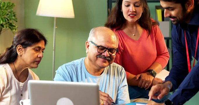 Senior Indian man signing document agreement as young executive guides him, seated beside wife and daughter in cozy modern home , symbolizing will, legal paperwork, finance or insurance consultation