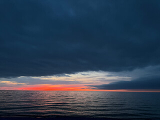 Obraz premium Cloudy sky over a calm sea at sunset with red and orange colors on the horizon. The majority of the sky is covered in dark clouds.