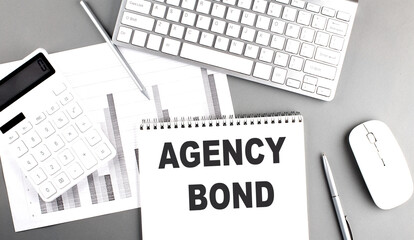 Agency bond phrase on spiral notepad with office supplies, charts, keyboard and calculator on gray desk background