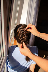 Naklejka premium Close-up of mother's hands weaving braid in daughter's hair. Beauty girly routine before school and feminine bonding. Authentic parenting moment.