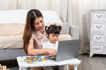 Asian woman early 30s sitting with toddler girl helping use laptop surrounded by colorful alphabet pieces on white table in cozy living room, concept mother daughter bonding early childhood education