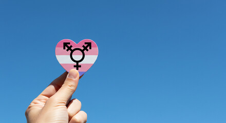 A hand holding a heart shaped transgender flag against a clear blue sky in bright sunlight outdoors