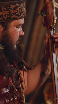 Bearded person plays two-string instrument while colleague taps drum set in traditional entertainment hub closeup. Concert of shamanic Altai music
