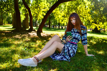 A beautiful lady in a summer dress is sitting on a lawn in a park, looking at her phone against the backdrop of trees.