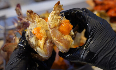 Close-up of black-gloved hands holding raw marinated crab halves with orange roe, a traditional...