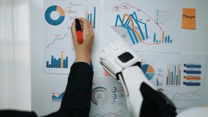A business professional utilizes a robotic arm to analyze various data charts, reflecting the integration of technology in modern workplaces for improved productivity and decision-making.Noogenesis