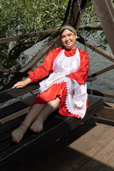A woman in a national Udmurt costume sits on a wooden swing by the water