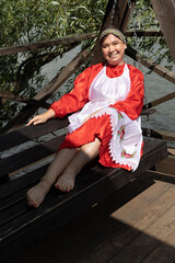 A woman in a national Udmurt costume sits on a wooden swing by the water