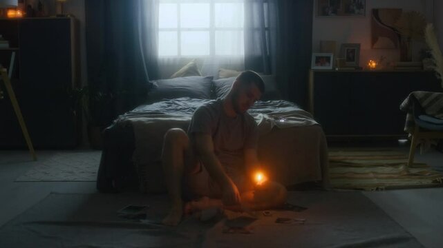 Full low key shot of young depressed melancholic man with broken heart from breakup sitting on floor amidst photos with his ex-love and burning one picture, feeling lonely and desperate