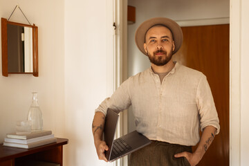 Portrait of a caucasian man with a beard at home in living room using a laptop wearing a hat and a linen shirt smiling. 