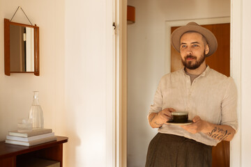 Portrait of a caucasian man with a beard at home in living room holding a coffee cup wearing a hat and a linen shirt smiling. 