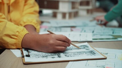 A young architect is focused on sketching building plans on paper with a pencil. The scene highlights the vibrant atmosphere of collaboration and creativity in a design workspace. SACTR