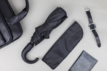 A black umbrella in a case lies on a table next to a man's watch and a leather bag