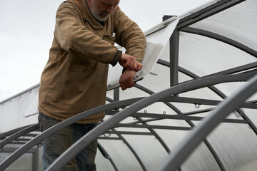 Installation of polycarbonate greenhouse. A man cuts off excess polycarbonate from the upper window sashes against the background of the metal frame.