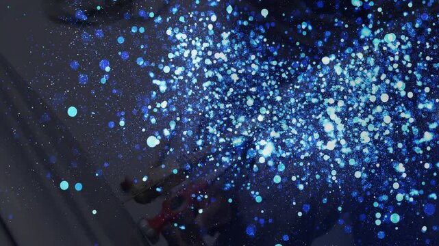 Technician gloved hand handling spray gun in industrial workshop, creating blue particle cloud