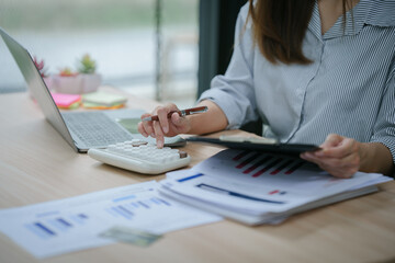 Young asian businesswoman holding pen and reading financial documents, smiling and working at office desk with computer showing graphs, startup business concept