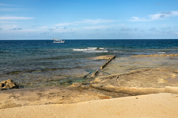 A seaside landscape with crystal-clear blue water, partially submerged natural rock formation, and a tourist boat on the horizon. A calm coastal scene with Mediterranean sunny weather.