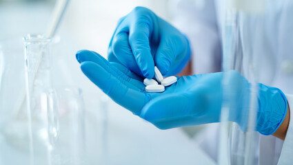 Close-up of hands wearing blue medical gloves carefully holding several white oval pills or tablets in a bright scientific laboratory setting with glassware