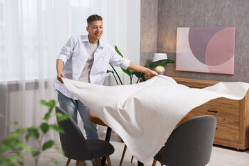 Young man putting white tablecloth on table at home
