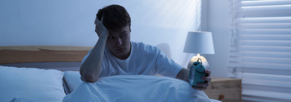 Frustrated man looking at alarm clock on bed at night. Insomnia problem