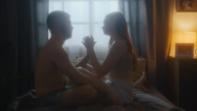 Side full shot of young affectionate couple touching each other hands and showing tenderness in bed in morning - stroking head and touching hair, looking in eyes with love
