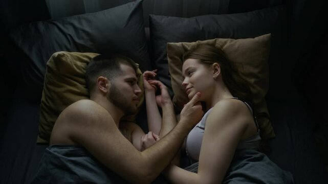 Top view of young man and woman in underwear lying in bed at home, facing each other, enjoying bond and connection. Gentle touches, subtle smiles, soft look in eyes, love in air, copy space
