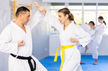 Obraz premium Adult man and young woman in kimono practicing karate techniques in group in gym..