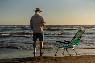 Senior man standing barefoot by the sea during sunset, holding a drink and gazing at the horizon, with an empty beach chair nearby on the shoreline.