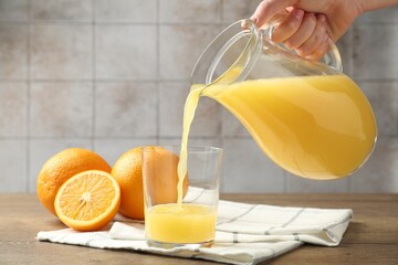 Woman pouring fresh orange juice from jug into glass at wooden table against grey tiled wall, closeup