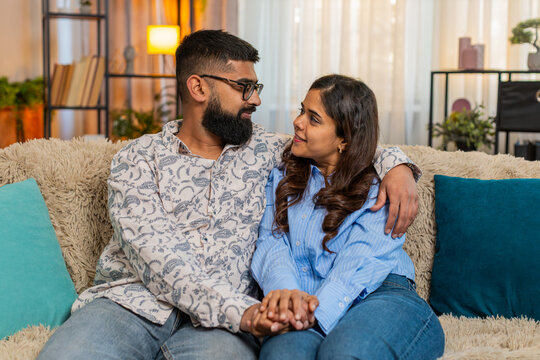 Indian couple man and woman sitting on home sofa, quietly holding hands and sharing peaceful emotional connection full of support. Young family enjoying silent harmonious moment with love and warmth